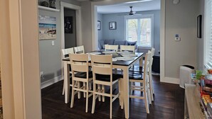 Uma sala de jantar com uma mesa posta para seis pessoas, cadeiras brancas, um ventilador de teto e uma janela com persianas.
