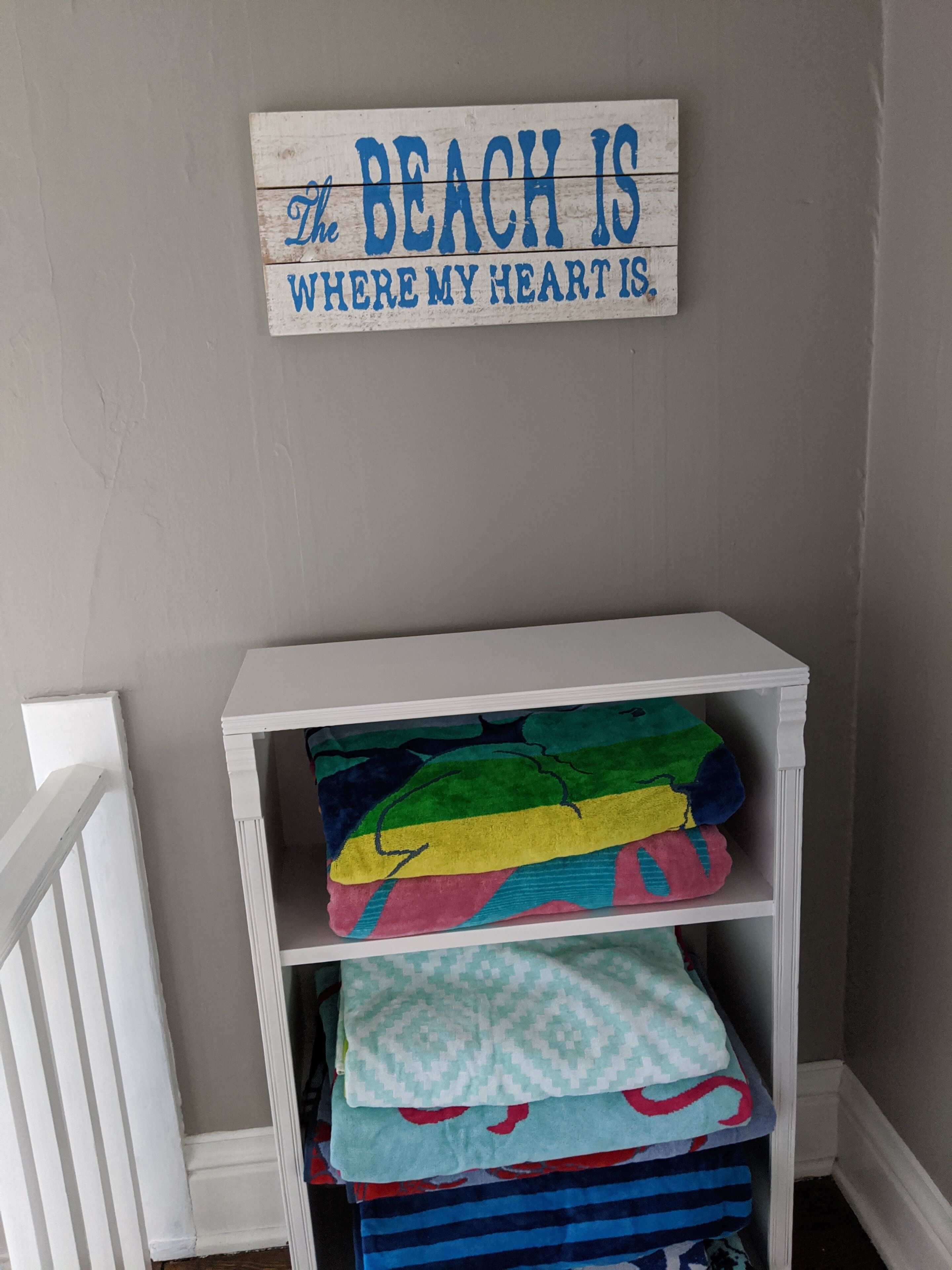 Un estante blanco con mantas coloridas y un cartel que dice 'The beach is where my heart is'.