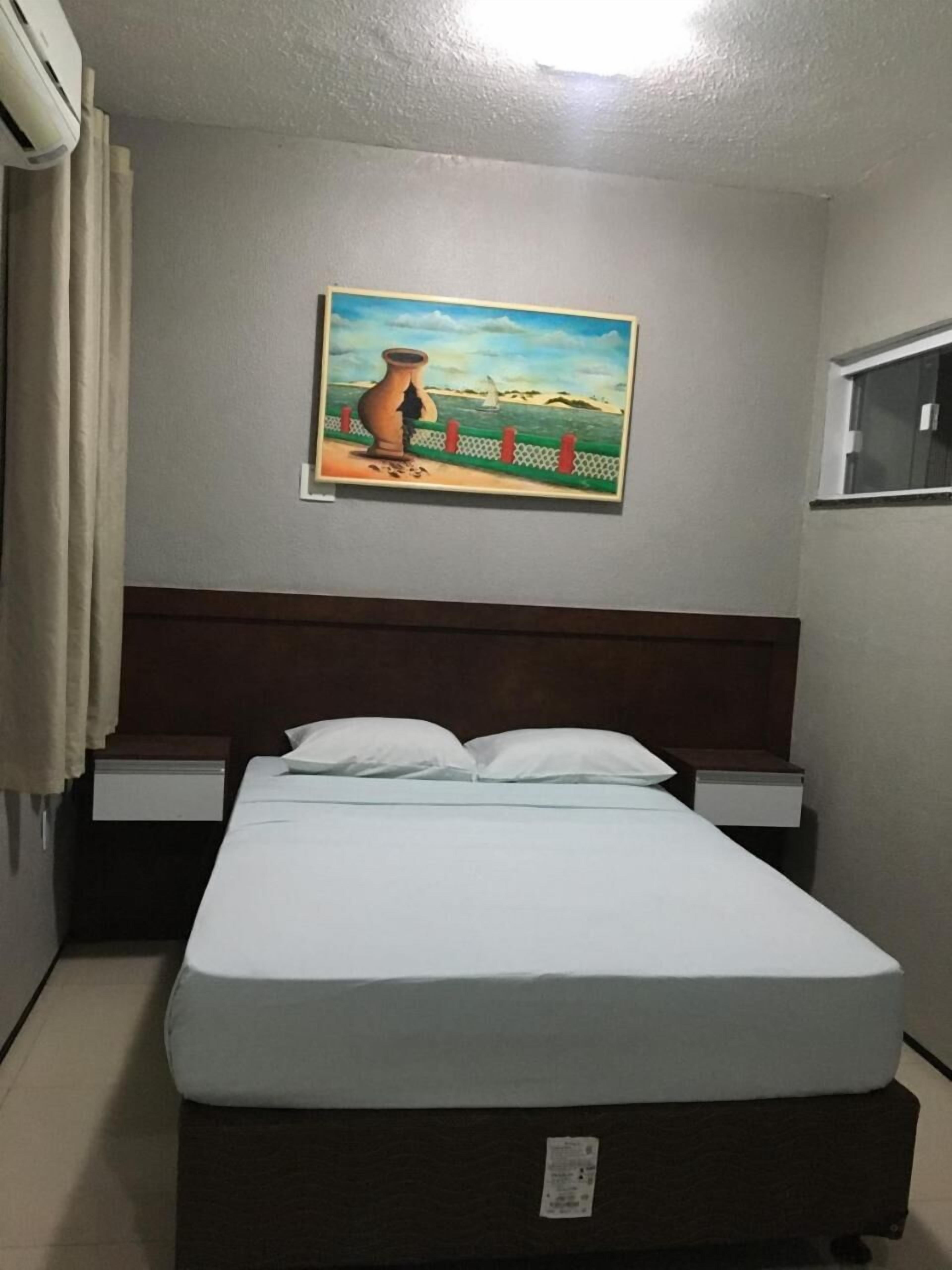 A hotel room with a bed, two bedside tables, a painting on the wall, and an air conditioning unit.