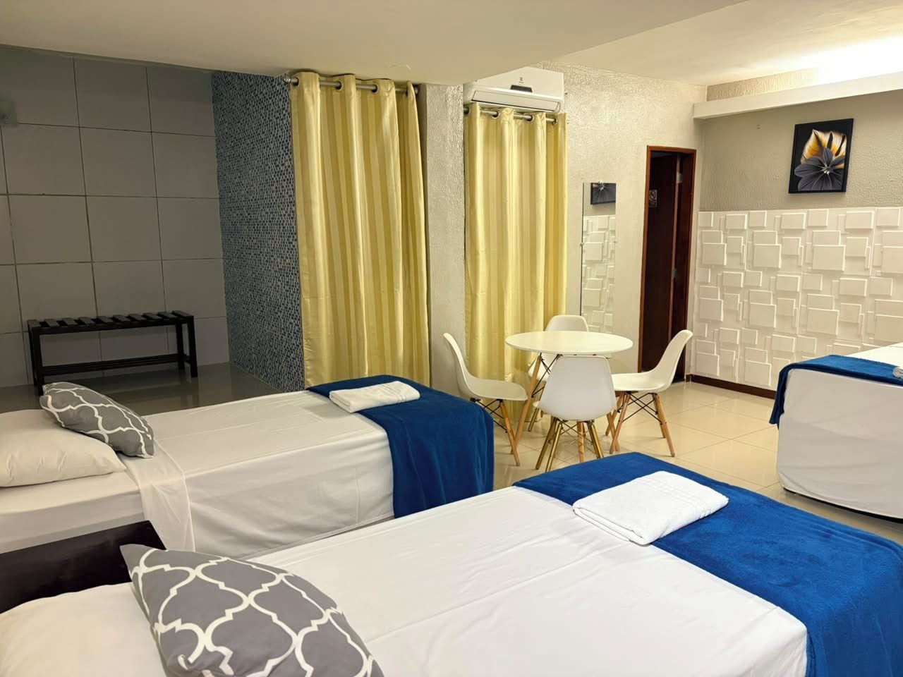 Habitación de hotel con dos camas, una mesita, sillas, un banco y un aire acondicionado instalado en la pared.