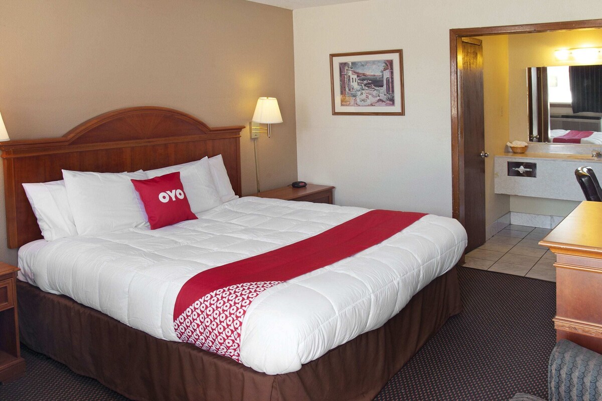 A hotel room with a large bed, a nightstand, a lamp, a framed picture on the wall, and a bathroom visible through an open door.