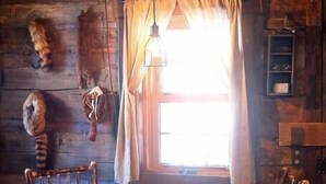 Ein rustikales Holzhaus-Interieur mit Esstisch und Stühlen, einer Pendelleuchte und einem Fenster mit Vorhängen.