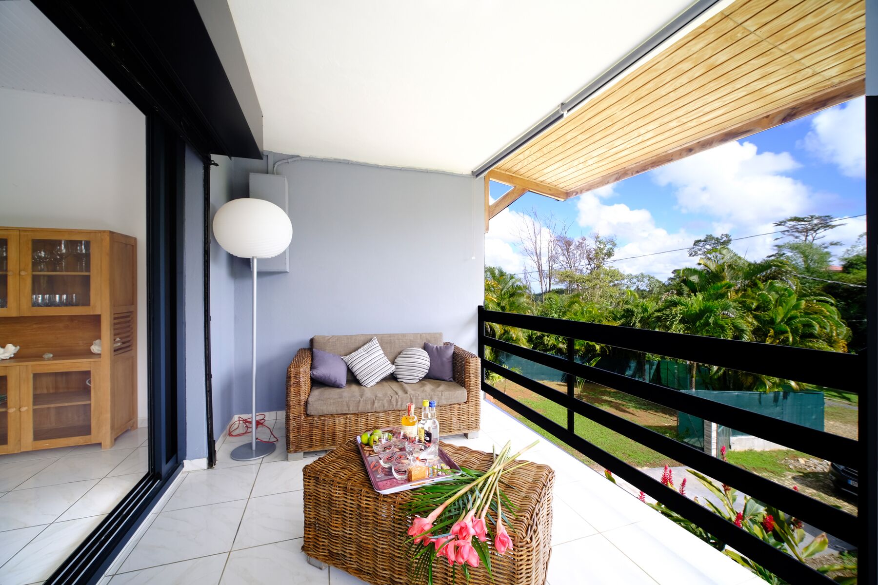 Un balcon avec un canapé en osier, une petite table d’appoint avec des boissons et des fleurs, et une vue sur la verdure.