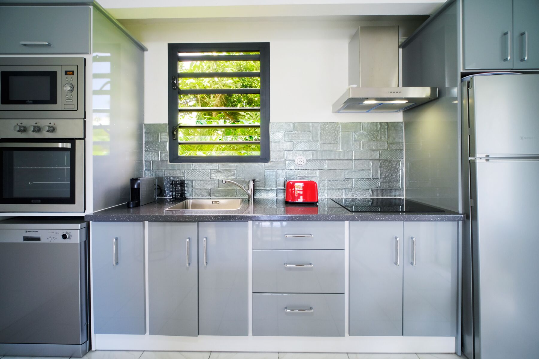 A modern kitchen with stainless steel appliances, grey cabinets, a window with a view of greenery, and a red toaster on the counter.