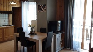 A dining area with a wooden table, chairs, a vase of flowers, a television on a cabinet, and a kitchen with wooden cabinets and a tiled backsplash. - Termignon Val cenis Vanoise (Termignon Val cenis)