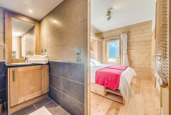 Une salle de bain moderne avec un grand miroir, des meubles en bois et un plan de travail noir. La pièce est dotée d’un plancher en bois et d’une grande fenêtre avec des rideaux.