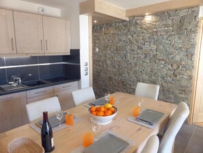 A modern kitchen with a stone wall, wooden cabinets, and a dining table set with orange fruits, glasses, and a bottle.