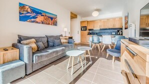 Ein modernes Wohnzimmer mit grauem Sofa, einem Couchtisch und einem Essbereich mit Stühlen. Im Hintergrund ist eine Küche zu sehen.