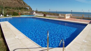 Ein rechteckiger Swimmingpool mit klarem, blauem Wasser, umgeben von einer gefliesten Terrasse. In der Nähe des Poolrandes stehen Liegestühle und ein Rettungsring. Auf der angrenzenden Straße ist ein Fahrzeug geparkt, und im Hintergrund ist ein Gebäude mit weißem Dach zu sehen.