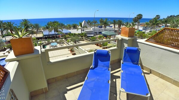 A rooftop terrace with two blue lounge chairs, a view of the ocean, and a palm tree garden. - 3 Bedrooms | Burriana Front Line | CG R924 (Nerja)