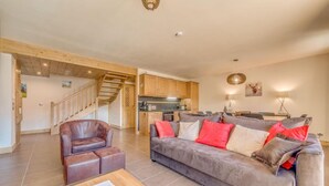 A spacious living room with a grey sofa, a brown leather chair, and a wooden coffee table. A kitchen is visible in the background with wooden cabinets and a staircase leading to another level. - 100m from resort center, 12nd floor, balcony, parking, tv, ski locker, 90m², Champagny-en-Vanoise (Champagny-en-Vanoise)