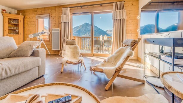 Un salon chaleureux avec un canapé, deux fauteuils recouverts de fourrure, un télescope et un balcon offrant une vue sur la montagne.