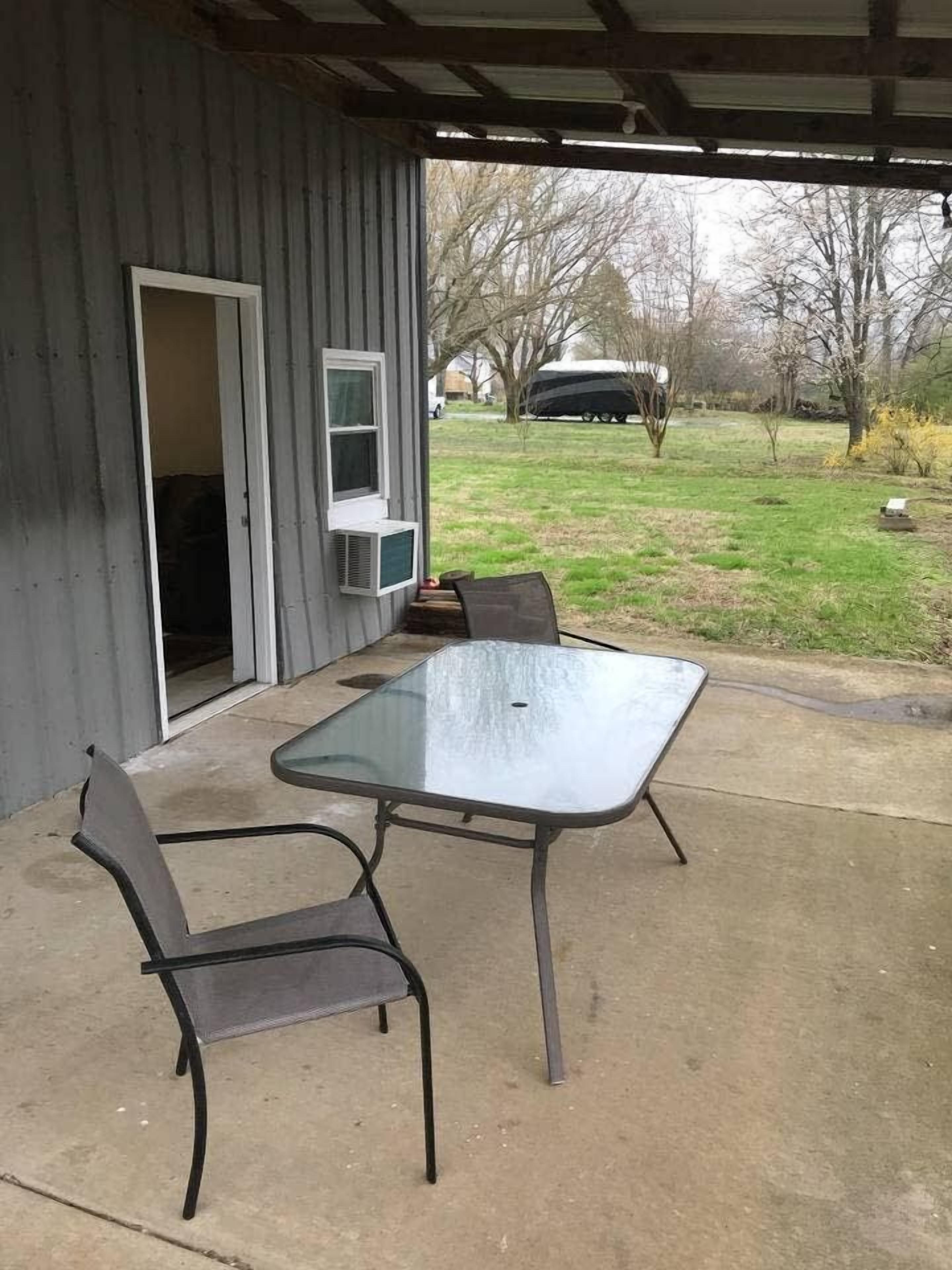 Une terrasse extérieure avec une table en verre et des chaises en métal, un abri couvert et une vue sur un véhicule et des arbres en arrière-plan.
