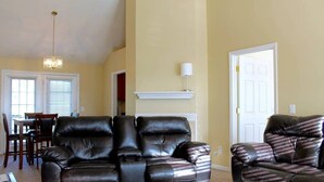Una sala de estar con un sofá de cuero oscuro, un comedor con mesa y sillas, y una puerta blanca.