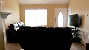 Una sala de estar con un sofá de color oscuro, un televisor sobre un mueble bajo y un amplio ventanal con persianas.
