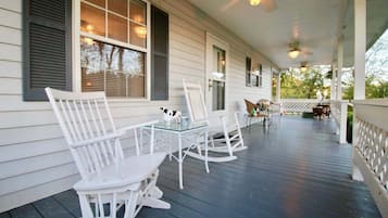 A covered porch with white rocking chairs and a glass table, featuring a ceiling fan and string lights.