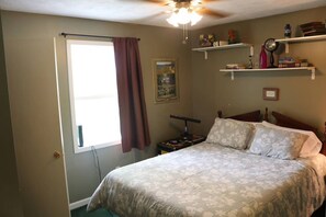 Une chambre à coucher avec un lit, un ventilateur de plafond, une fenêtre avec un rideau et des étagères remplies de livres et d’objets décoratifs.
