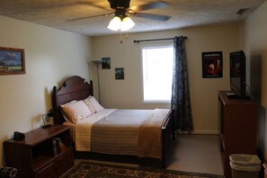 A bedroom with a bed, a ceiling fan, a window with curtains, a bedside table, a lamp, a dresser, and a poster on the wall.