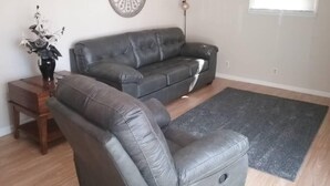 Ett vardagsrum med en grå lädersoffa, en tillhörande fåtölj, ett träbord med en vas, en dekorativ väggdekoration och en golvlampa.