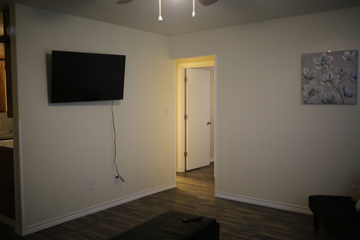 A room with a flat-screen TV mounted on the wall, a wooden floor, and a door leading to another room.