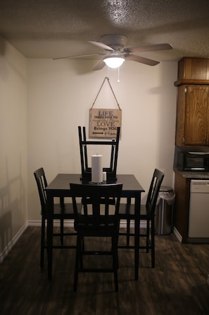 Uma área de refeições com mesa, cadeiras, ventilador de teto, uma placa pendurada e um micro-ondas ao fundo.