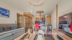 Un salon moderne avec un canapé, une table basse et un téléviseur. On aperçoit un coin repas avec des chaises et, au fond, une cuisine.
