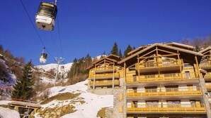 Un télésiège avec une cabine, des bâtiments de style chalet en bois dotés de balcons, et un paysage montagneux enneigé en arrière-plan.