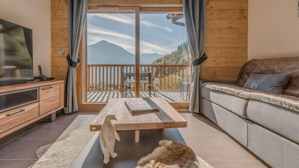 Un salon chaleureux avec un plancher en bois, un canapé, un téléviseur, une table en bois et une vue sur les montagnes par une porte vitrée.