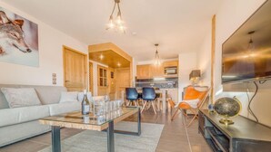 Una sala moderna con sofá, mesa de centro y sillones. Hay una zona de cocina con armarios de madera y una mesa de comedor. Un televisor está montado en la pared, y un globo se encuentra sobre una estantería.