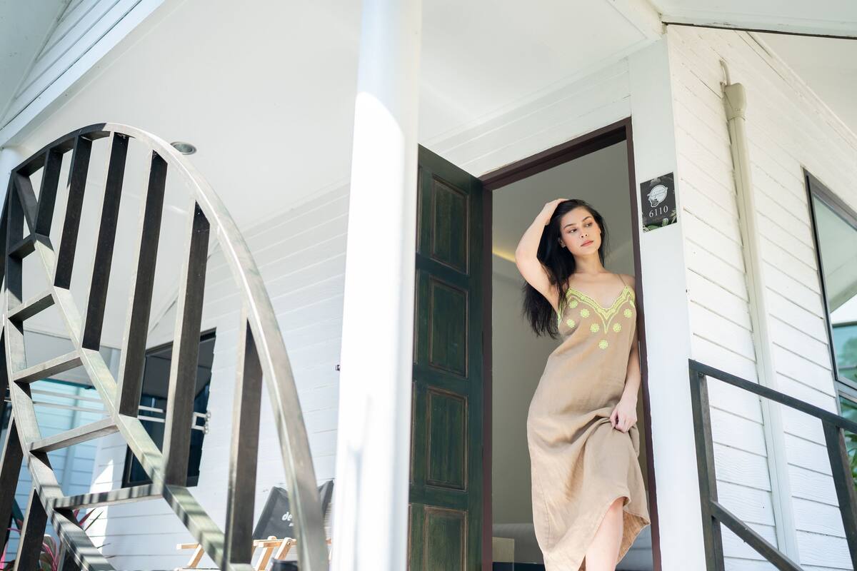 Una mujer con un vestido color beige está parada frente a la entrada de una casa con puerta verde y fachada blanca.