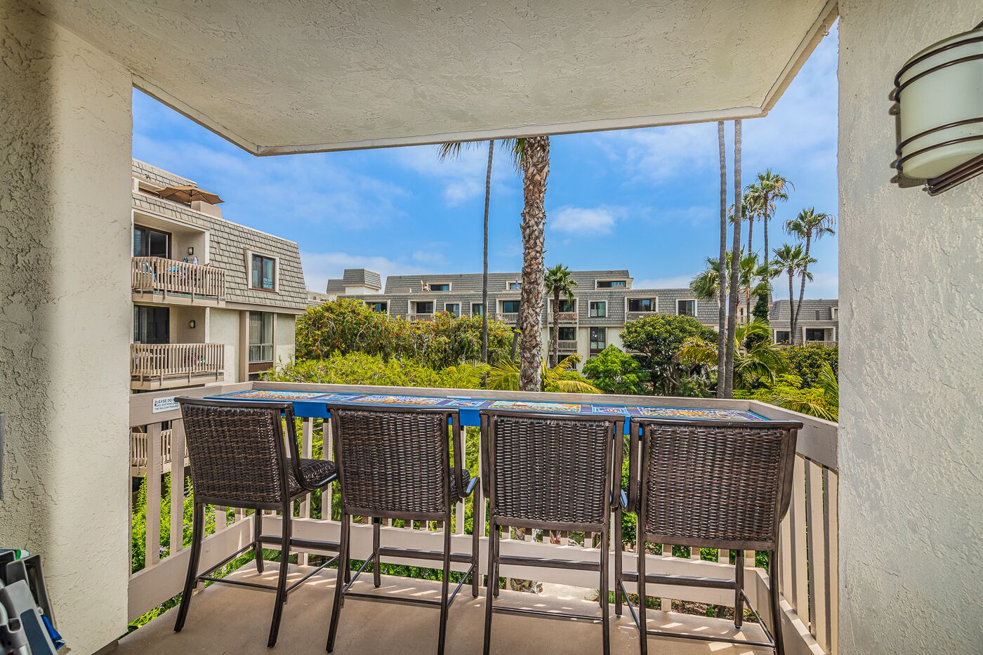 Un espacio al aire libre techado con una mesa y sillas, con vistas a un conjunto residencial con múltiples balcones y palmeras.