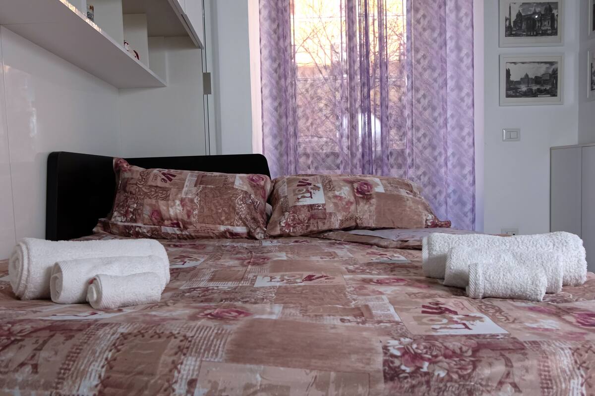 A neatly made bed with floral bedding, a headboard, and rolled white towels.