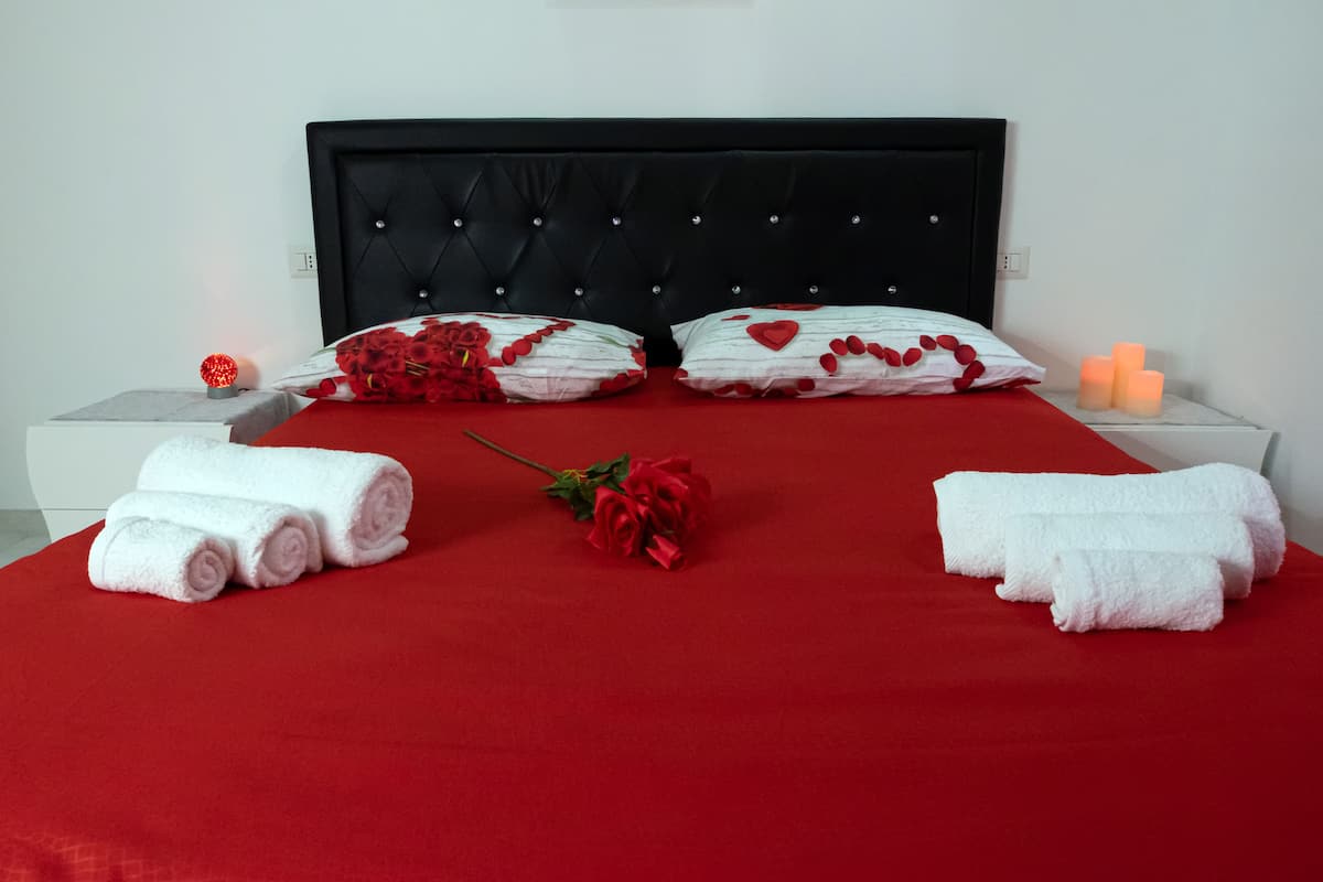 A neatly made bed with red and white linens, a bouquet of red roses, and white towels on a red bedspread.