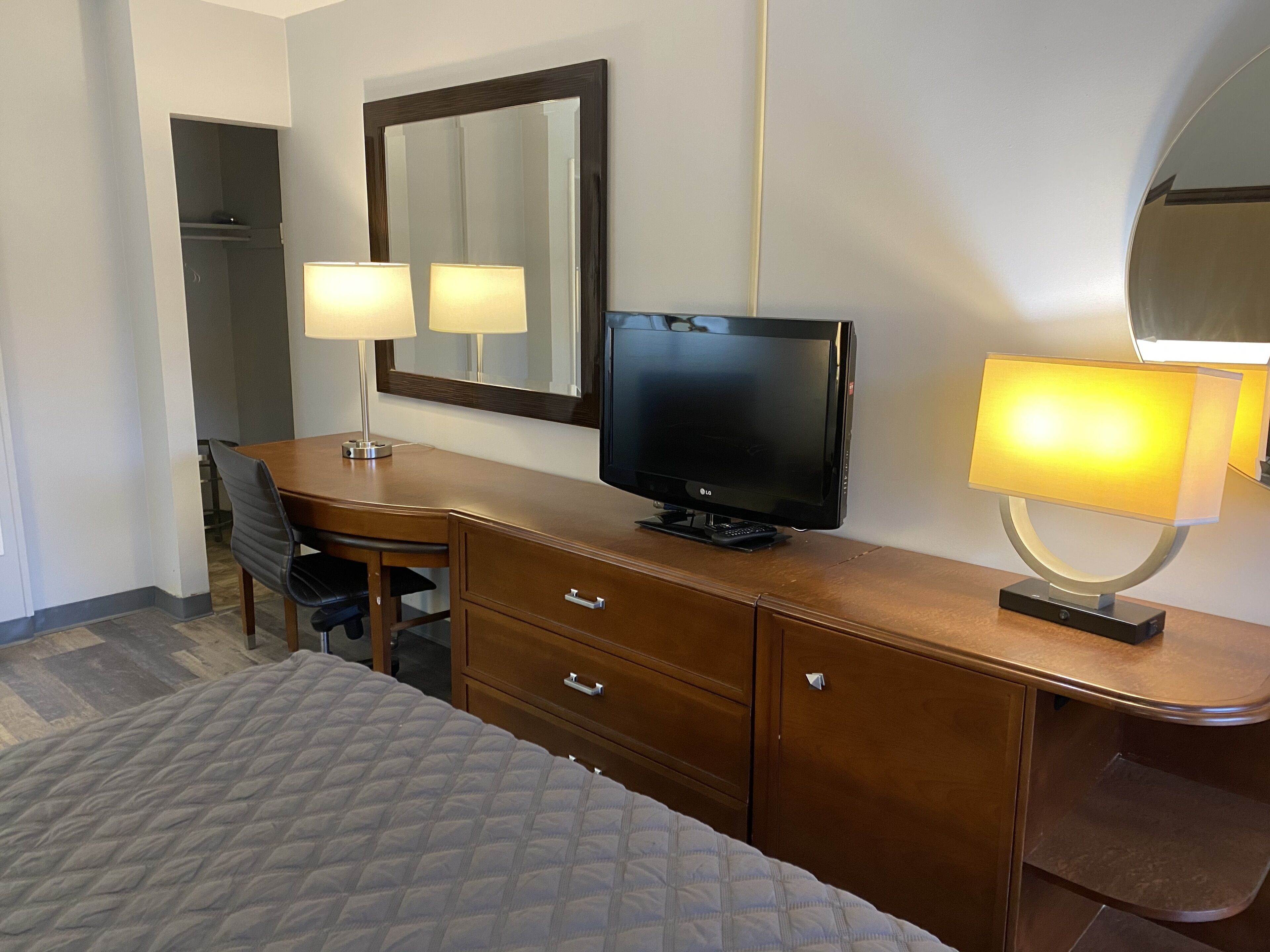 Una habitación de hotel con una cama, un escritorio de madera con televisor, dos lámparas y un espejo.