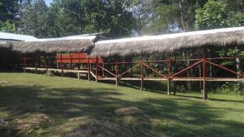 A thatched-roof structure with a wooden frame, surrounded by a fenced area and trees.