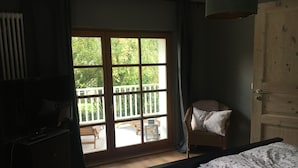 Quarto com cama, uma cadeira, televisão e porta de vidro deslizante que dá para área externa com mesa e cadeiras.