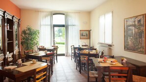 A dining area with multiple wooden tables and chairs, a large window with curtains, a radiator, and a painting on the wall. - Agriturismo Il Mondo (Parma)