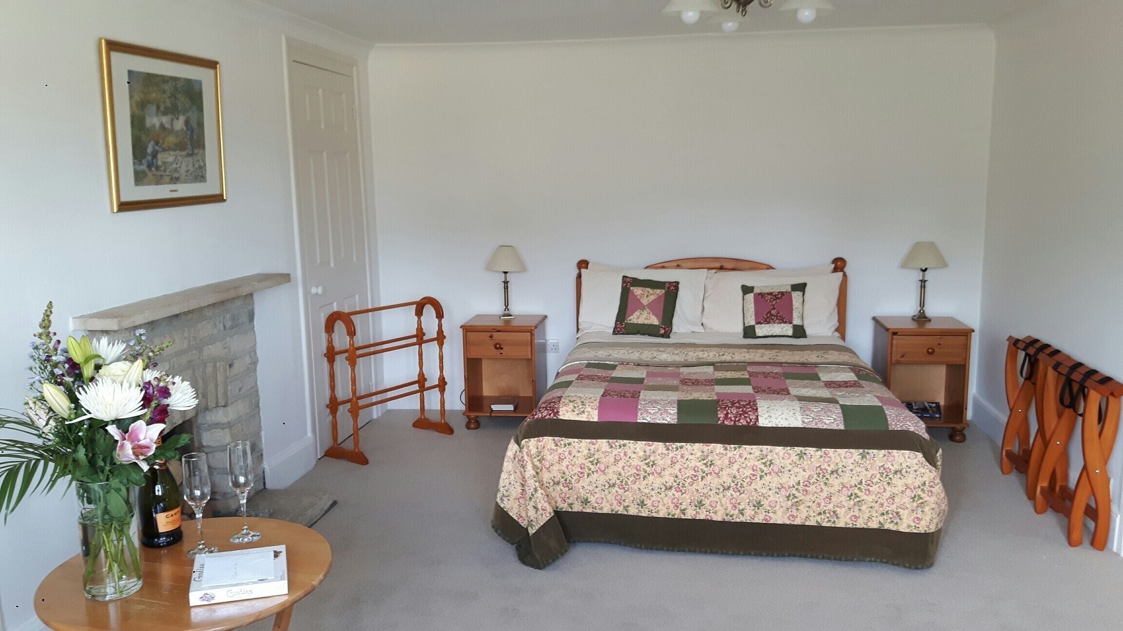 A bedroom with a bed, two bedside tables with lamps, a wooden chair, a small table with a vase and wine glasses, and a painting on the wall.