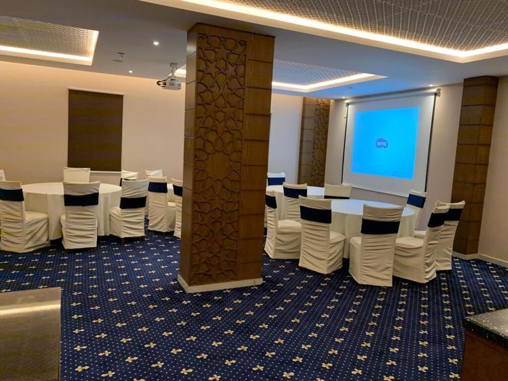 Une salle de conférence équipée de tables rondes et de chaises, d’un écran de projection et d’un tapis à motifs.