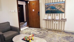 Un vestíbulo de hotel con mostrador de recepción, un televisor mostrando un paisaje urbano, una zona de estar con una mesa y una puerta marcada con un número de habitación.