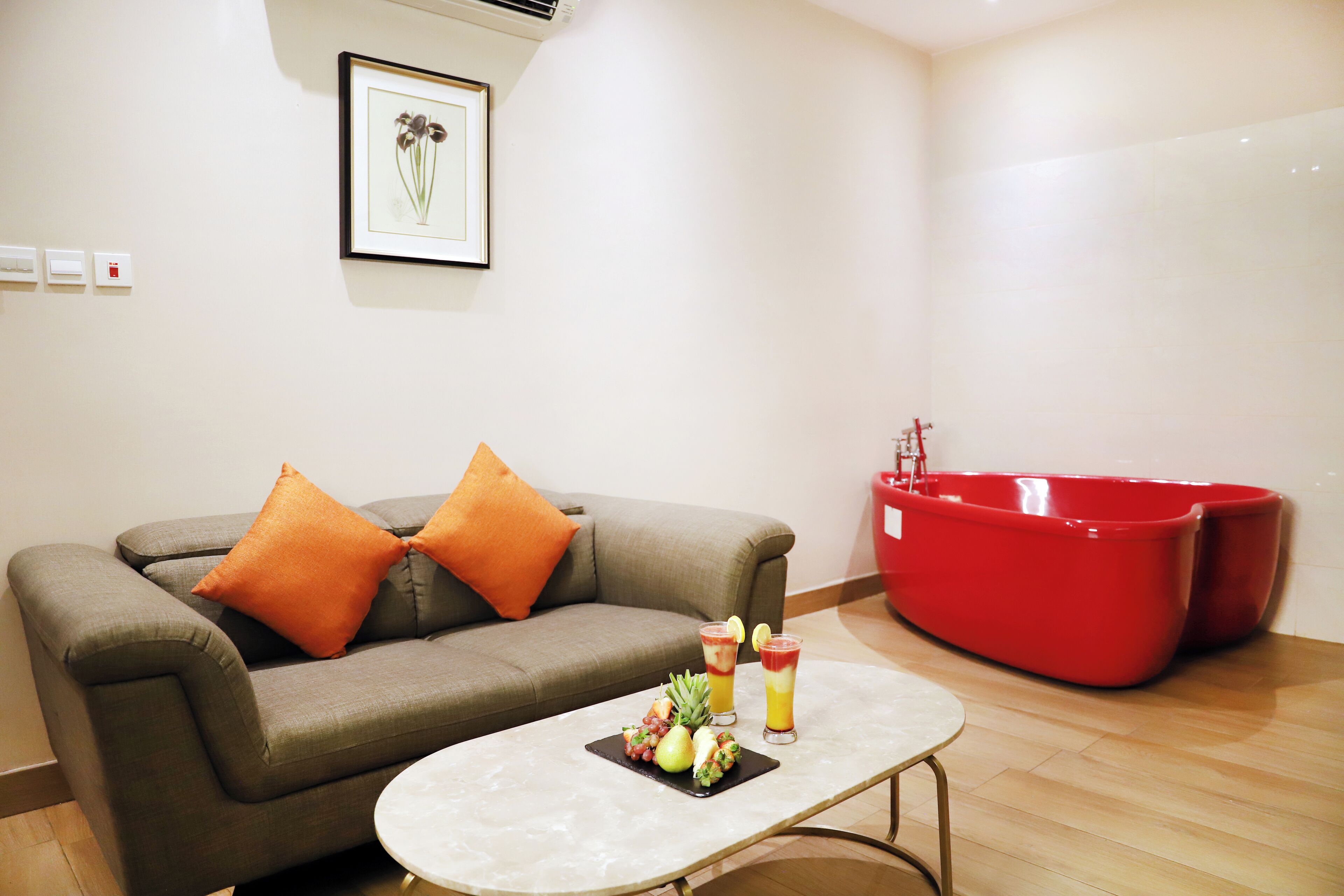 A modern living room with a grey sofa, orange cushions, a marble coffee table with fruit and drinks, and a red bathtub.
