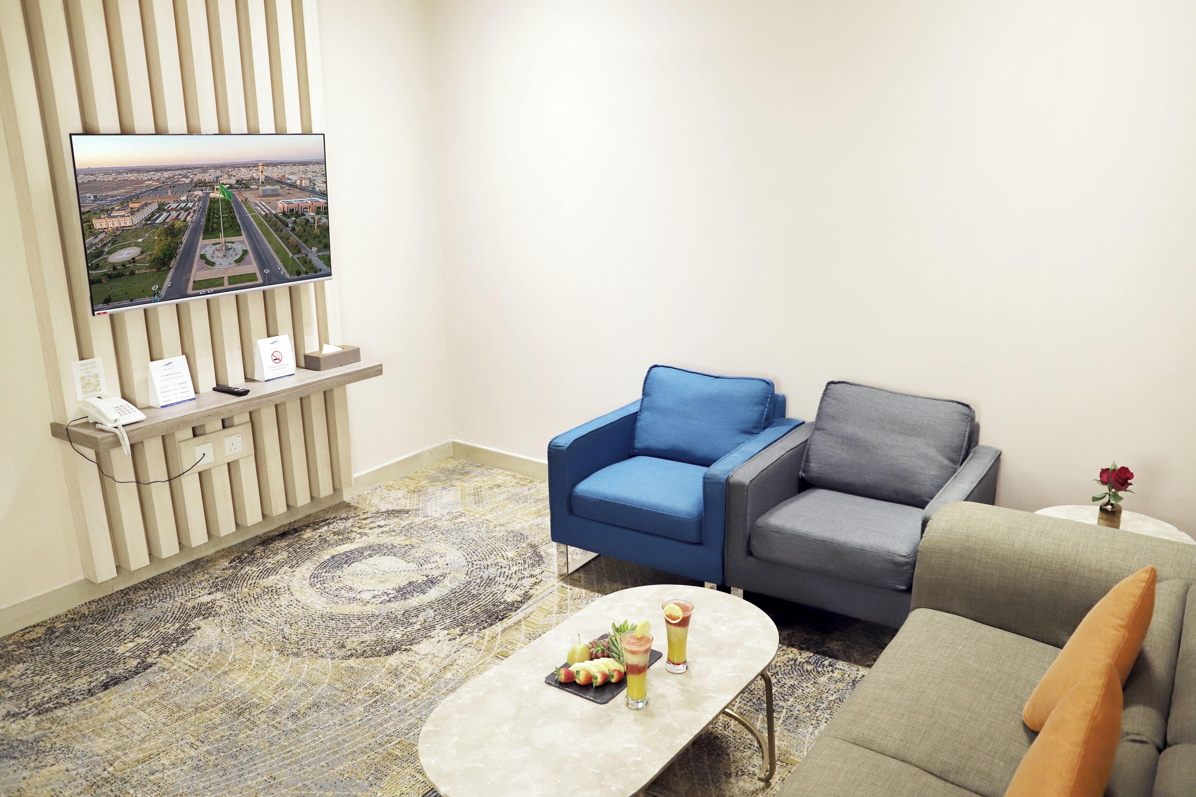 A modern living room with a flat-screen TV mounted on a wall, two sofas, a coffee table with fruit and drinks, and a patterned carpet.
