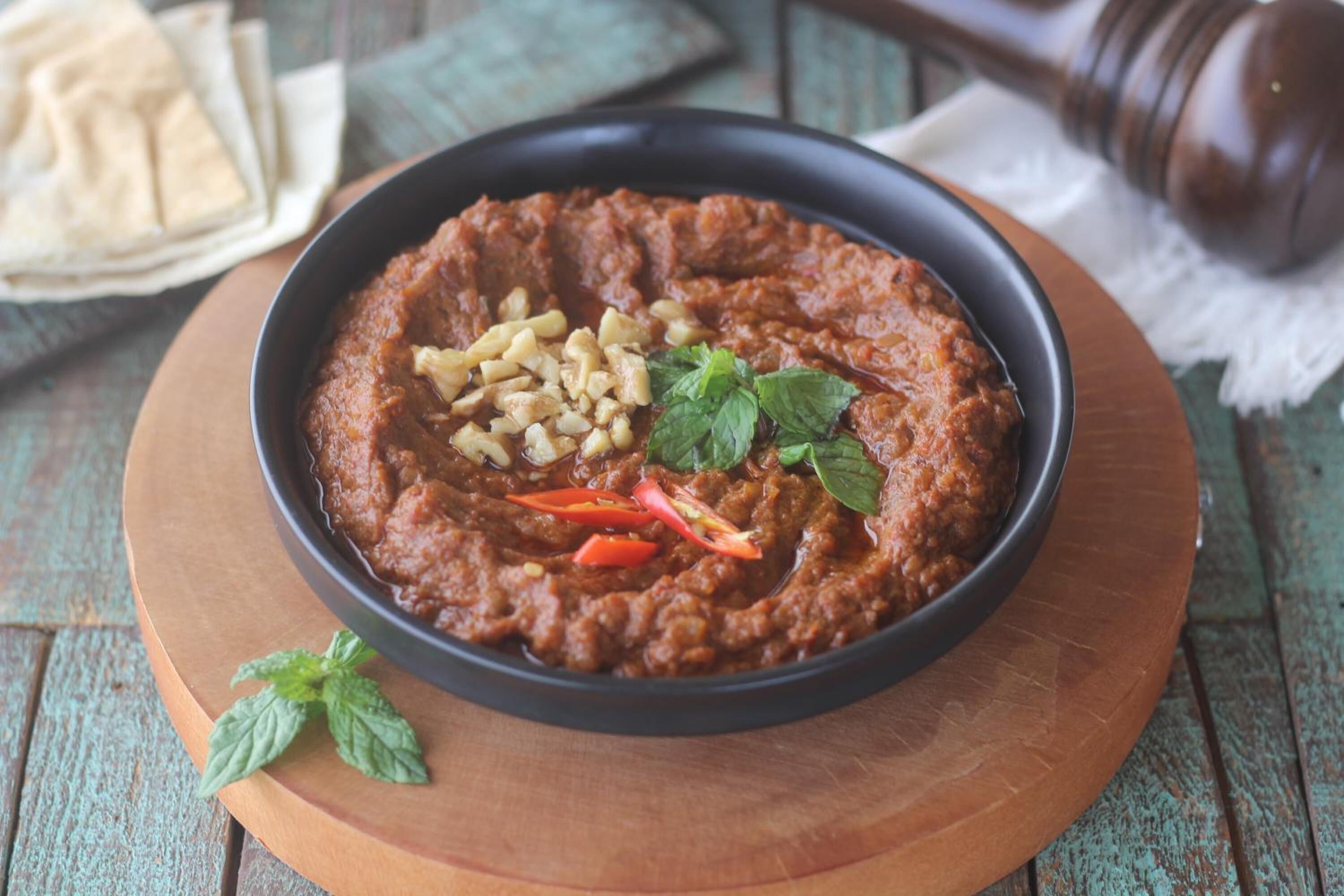 A bowl of hummus garnished with pine nuts and red chili slices, accompanied by a folded piece of flatbread and a mint leaf on a wooden board.