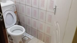 Une petite salle de bain avec des toilettes, une douche et un lavabo. Les murs sont recouverts de carreaux disposés selon un motif, et il y a un rouleau de papier toilette sur les toilettes.