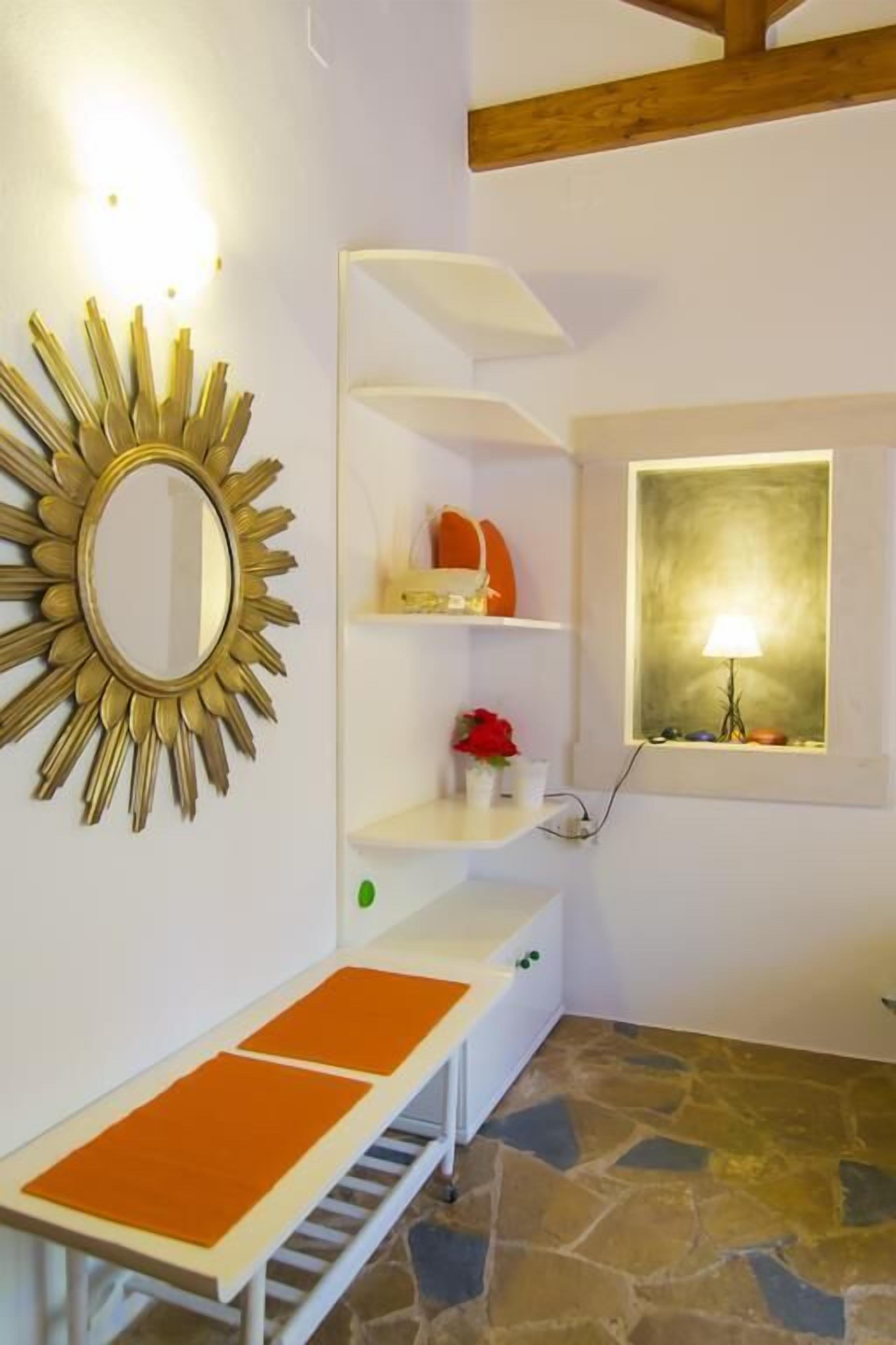 Um quarto com espelho em formato de sol, estante branca, pequena mesa com almofadas laranja e janela com luminária.