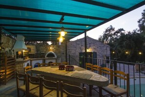 Un espace extérieur aménagé pour les repas, avec une table et des chaises en pierre, un four à pizza et une terrasse couverte avec un toit bleu.