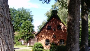 Ein rustikales Backsteingebäude mit geneigtem Dach, umgeben von üppiger Vegetation und einem Holzzaun.