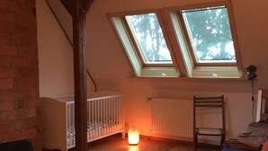 Ein Zimmer mit schrägem Dach, zwei Dachfenstern, einem Babybett, einem Stuhl und einem Bett.