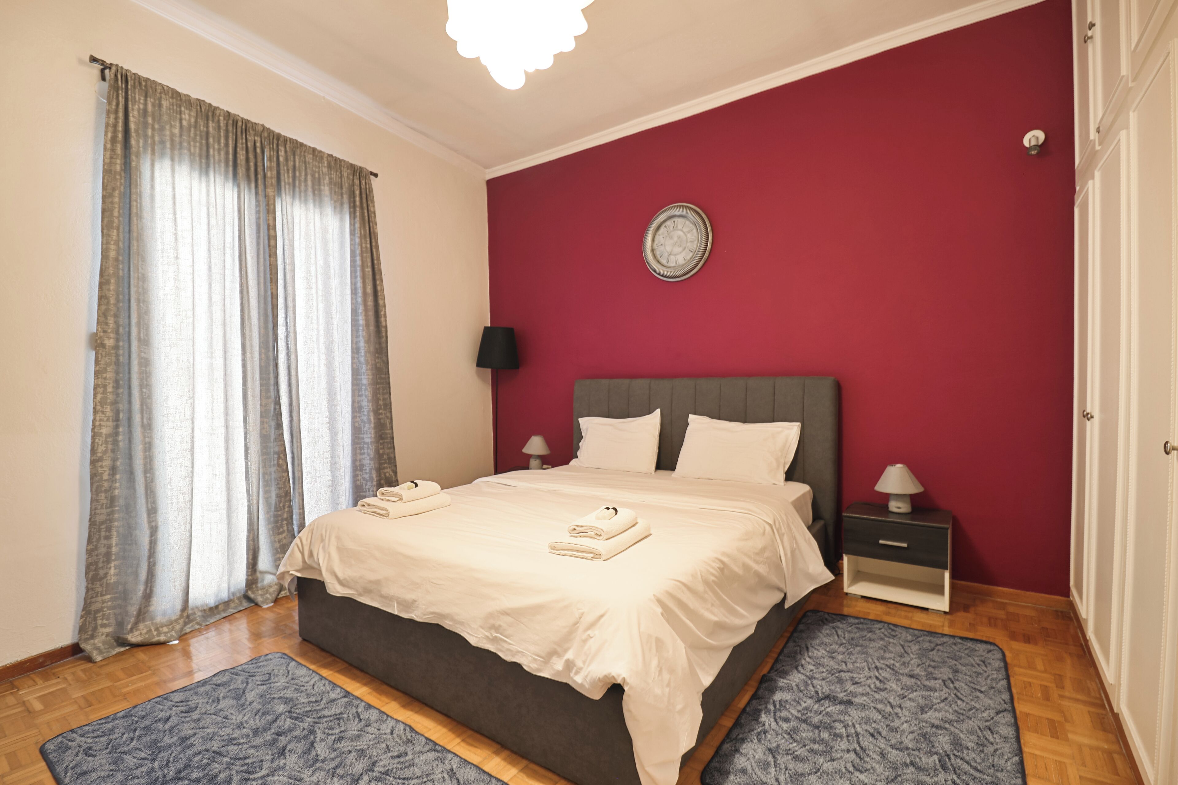 Un dormitorio con una cama, dos mesitas de noche, una mesita auxiliar, un reloj de pared y una ventana con cortinas.