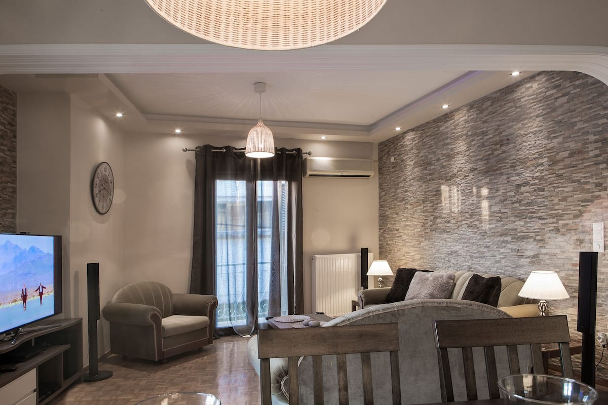 A modern living room with a large flat-screen TV, comfortable seating, a dining area, and a stone-textured wall.